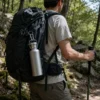 A TiRoam pure titanium water bottle clipped to a hiker’s backpack on a forest trail, showing a lightweight carry setup for backpacking, hiking, and travel.