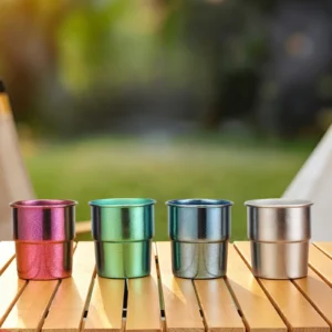Four colorful TiRoam anodized titanium stackable cups in purple, green, blue, and silver lined up on a wooden camping table.
