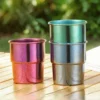Three TiRoam colorful titanium camp cups on a wooden outdoor table, showing one green cup stacked on a blue cup next to a purple cup.