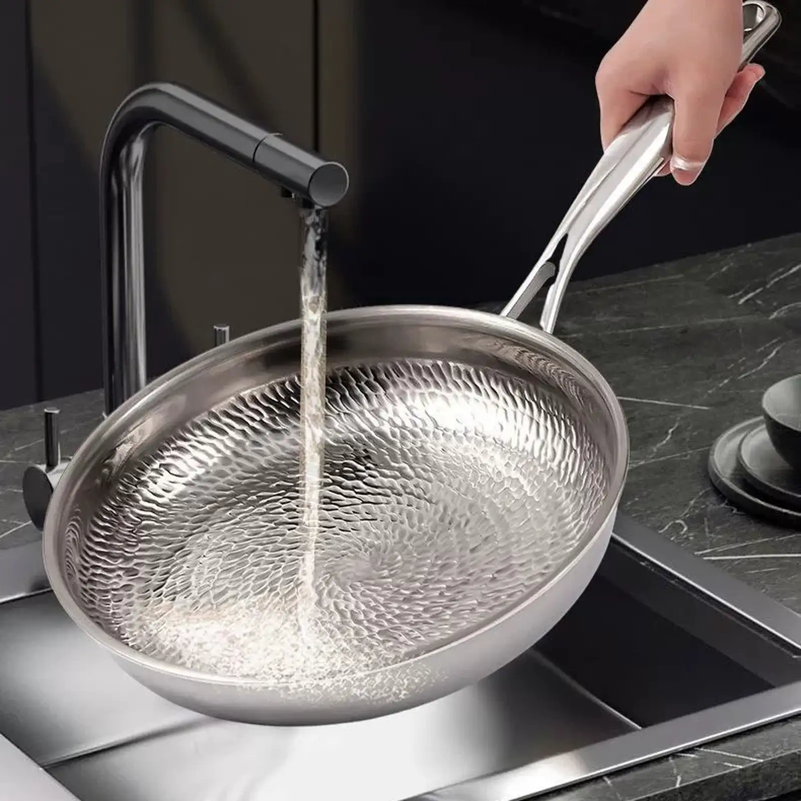 Water rinsing the textured cooking surface of a titanium frying pan under a kitchen faucet.